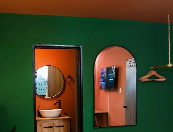 A forest-green wall with a doorway into a terracotta bathroom with a round mirror and vessel sink, beside a tall arched mirror reflecting a wall-mounted TV.