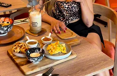 A wooden board at Canela Café with scrambled eggs, small waffles, dipping sauces and a fruit bowl, a bottled drink behind; a guest sits at the table, face out of frame.