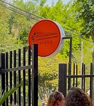 The round orange Canela Café sign on Bohemia’s black entrance gate, palm leaves and trees behind.