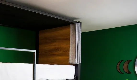 Black-framed capsule bunks with a warm wood panel and a grey privacy curtain against a deep green wall decorated with wooden crescent moons.