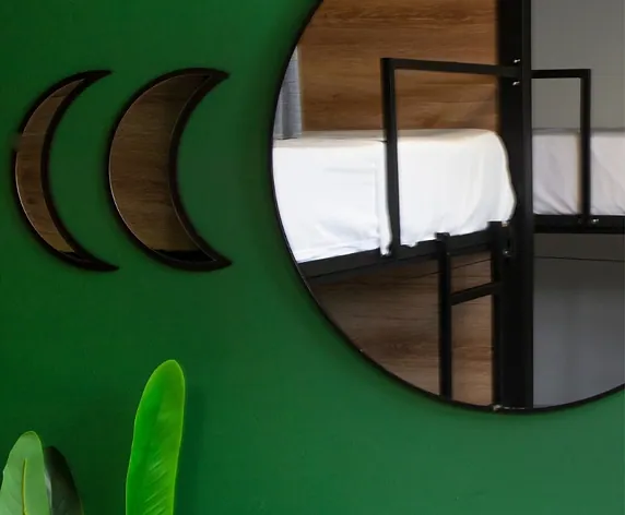 Two wooden crescent moons and a large round mirror on a green dorm wall; the mirror reflects a black bunk bed with white sheets.
