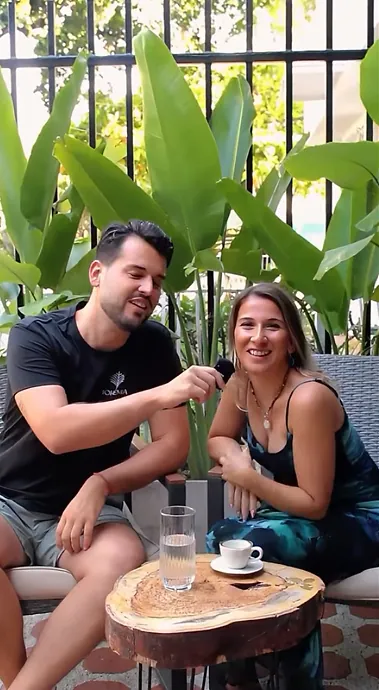 Marco and Ceren, Bohemia’s owners, laughing on a woven sofa among big banana-leaf plants, a coffee and a glass of water on a wooden slab table.