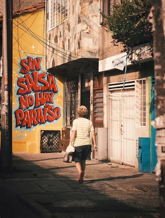 A yellow wall painted with huge red letters reading “Sin salsa no hay paraíso” on a sunny Cali street, an older woman walking past with a shopping bag.