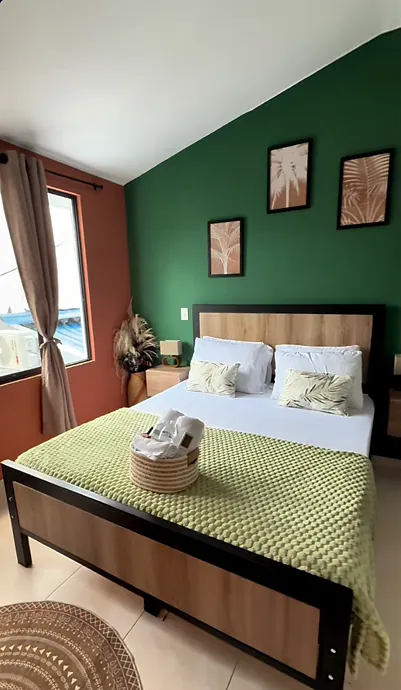 A wide wooden bed with an olive knit throw and a towel basket under a deep green accent wall hung with three botanical palm prints, a terracotta wall and curtained window to the left.